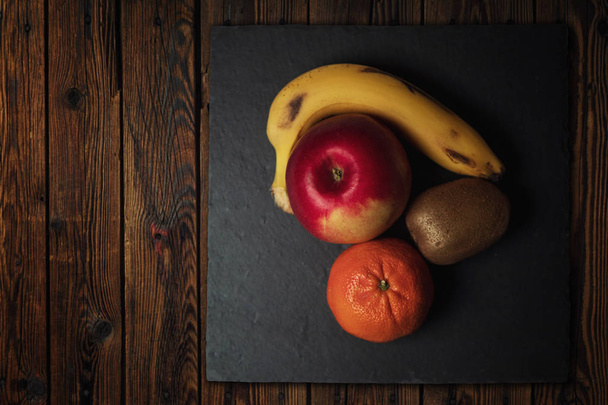 ganze Früchte auf einem schwarzen Teller auf einem Holzbrett. Banane, Apfel, Kiwi, Mandarine. Blick von oben - Foto, Bild