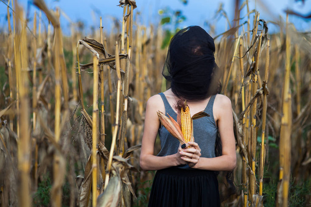 a strange portrait of a girl without a face with a black cloth on her head on a dried corn field holds an ear of corn at her chest - Foto, Bild