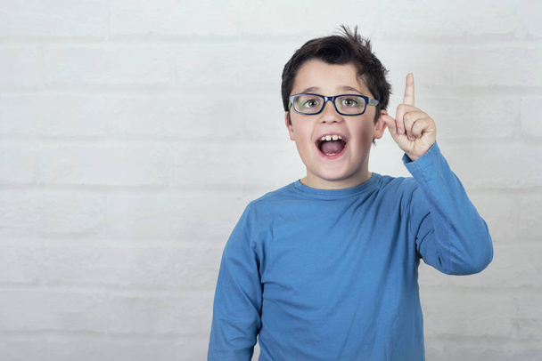 мальчик в очках поднимает руку, указывая указательным пальцем на кирпичный фон
 - Фото, изображение