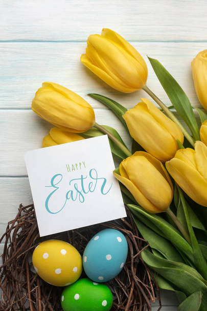 Velikonoční pozadí světlé, barevné vejce v hnízdě a žluté tulipány. Pohled shora s prostorem pro vaše pozdravy - Fotografie, Obrázek