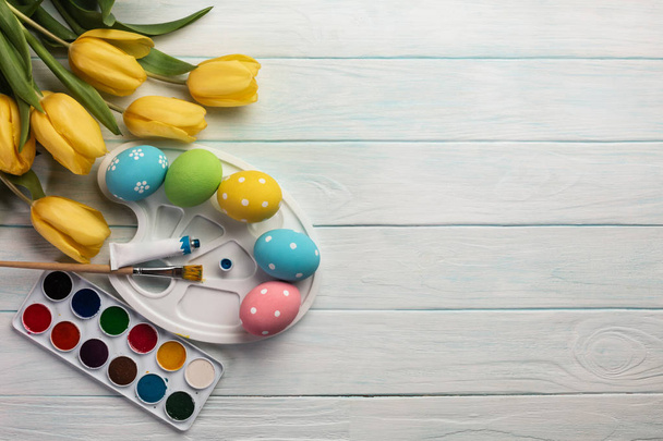 Ovos de páscoa coloridos, paleta de tinta, pincéis e tulipas amarelas na mesa de madeira branca. Vista superior com espaço de cópia
 - Foto, Imagem