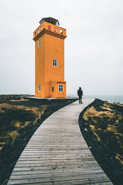 Unidentified Person standing in front of Orange Lighthouse Svortuloft Skalasnagi tower in Snaefellsnes Peninsula, west Iceland on an overcast day. - 写真・画像
