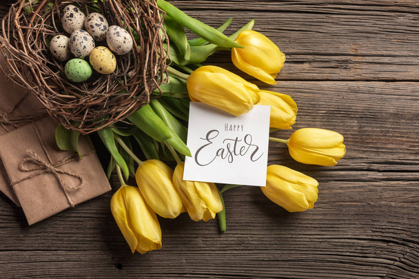 Tulipanes amarillos en una bolsa de papel, un nido con huevos de Pascua sobre un fondo de madera. Vista superior con espacio de copia
 - Foto, Imagen