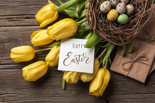 Žluté tulipány v papírovém sáčku, hnízdo s velikonoční vejce na dřevěné pozadí. Pohled shora s kopií prostor - Fotografie, Obrázek