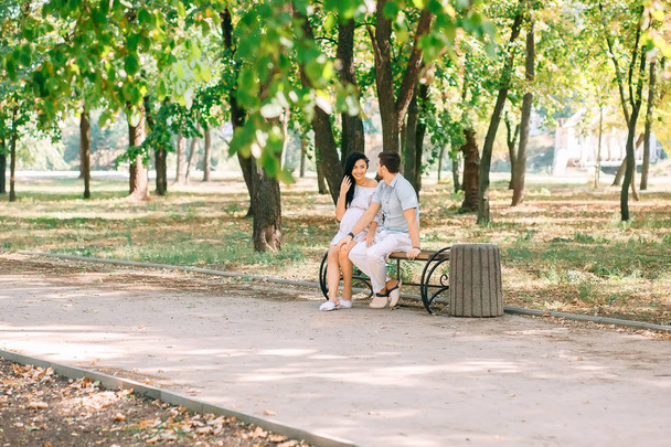 A young guy and his pregnant girl are sitting on a bench, against the background of greenery and trees. In anticipation of a child. First pregnancy I love children. - Photo, image