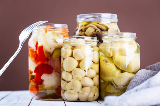 Marinated mushrooms, artichokes and various vegetables in a glass jars on white wooden background. Selective focus  - Foto, imagen