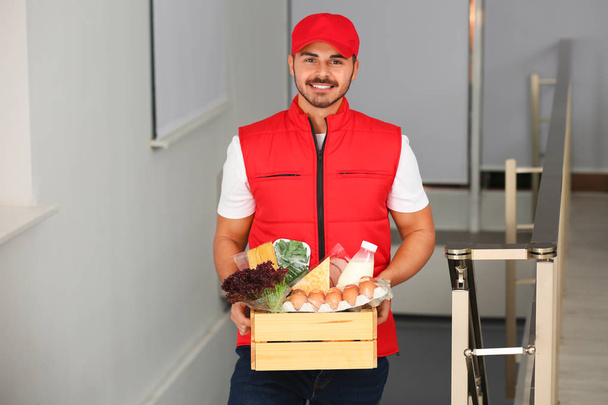 Male courier holding wooden crate with products indoors. Food delivery service - Foto, Imagem