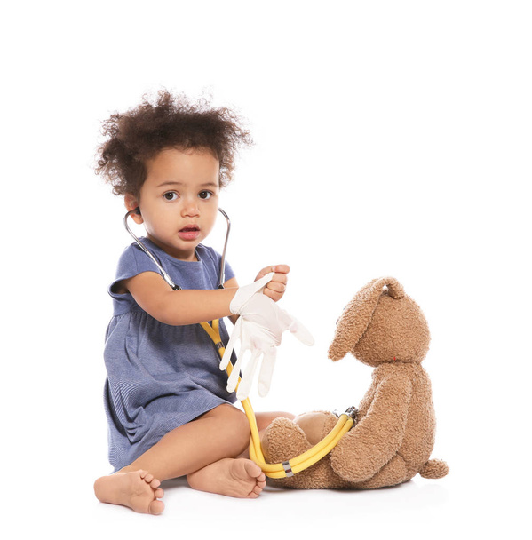 Cute African American child imagining herself as doctor while playing with stethoscope and toy bunny on white background - Photo, Image
