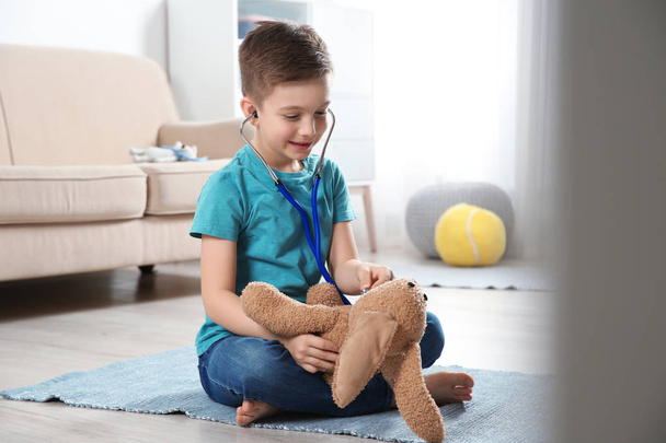 Cute child imagining himself as doctor while playing with stethoscope and toy bunny at home - Φωτογραφία, εικόνα