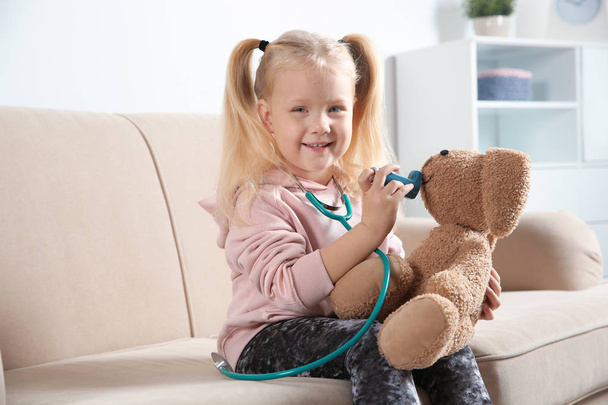 Cute child imagining herself as doctor while playing with toy bunny on couch at home - Valokuva, kuva