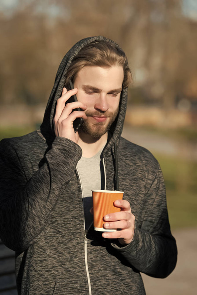 New technology and modern life. Guy with smartphone and takeaway drink on fresh air. Bearded man in hood hold disposable coffee cup on sunny outdoor. Coffee or tea mood. Morning tea for breakfast - Valokuva, kuva