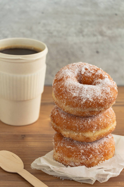 A stack of sugar coated doughnuts on crumpled paper with a hot cup of coffee , on wooden background. - Photo, Image