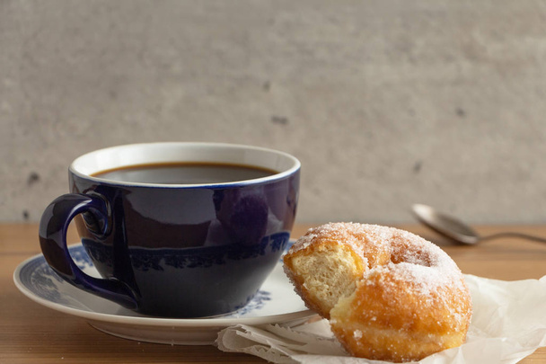 zuckerbeschichteter Donut auf zerknülltem Papier mit einer Tasse Kaffee, auf hölzerner Tischplatte. - Foto, Bild