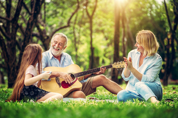 Счастливая семья играет на гитаре и поет вместе, сидя в парке летом. Концепция семейной связи по музыке
. - Фото, изображение