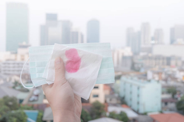 Woman hands or female hands holding a protective mask and napkin with blood because of air pollution in the city have particulate matters or PM 2.5 with city building background - Photo, Image