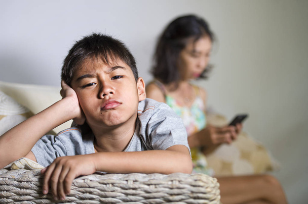 νέοι λυπημένος και βαριούνται 7 ή 8 χρόνια παλιά Ασίας παιδί στο σπίτι καναπέ αισθάνονται απογοητευμένοι και χωρίς παρακολούθηση ενώ μητέρα δικτύωσης στο κινητό τηλέφωνο ως internet εξαρτημένος παραμελεί το γιο της  - Φωτογραφία, εικόνα
