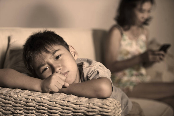 young sad and bored 7 or 8 years old Asian child at home couch feeling frustrated and unattended while mother networking on mobile phone as internet addict neglecting her son  - 写真・画像
