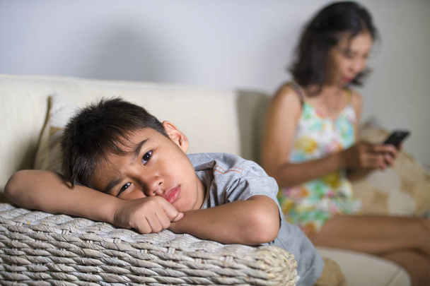 young sad and bored 7 or 8 years old Asian child at home couch feeling frustrated and unattended while mother networking on mobile phone as internet addict neglecting her son  - 写真・画像
