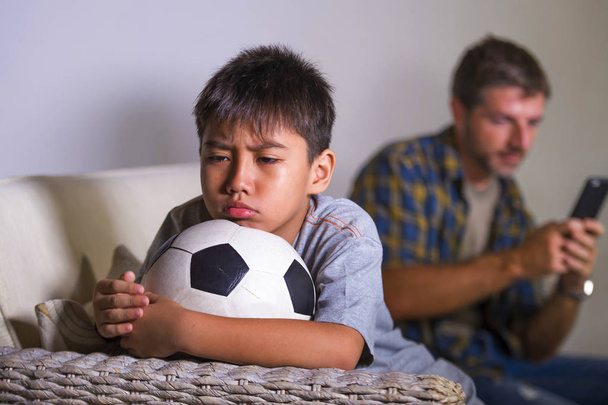 young sad and bored child at home couch feeling frustrated and unattended waiting his father for playing football while man networking on mobile phone as internet addict father neglecting son - Zdjęcie, obraz