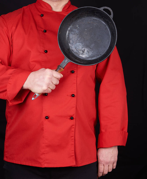 cook in red uniform holding an empty round black frying pan, black background - Foto, Imagen