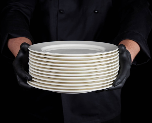 μάγειρας με μαύρη στολή και μαύρα γάντια λατέξ κρατά στα χέρια του μια στοίβα από στρογγυλά λευκά άδεια πιάτα - Φωτογραφία, εικόνα