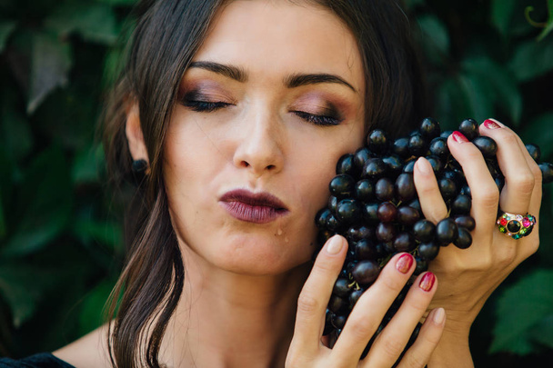 Portrait of young sexy brunette in black dress biting black grapes. - Foto, Imagem