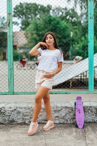 Summer lifestyle image of trendy pretty young girl standing next to the scateboard coart with her purple plastic scateboard. - Foto, afbeelding