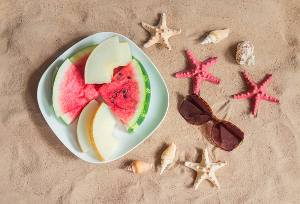 Piatto con fette di melone e anguria, occhiali da sole, stelle marine e conchiglie sulla spiaggia di sabbia. Vista dall'alto. Concetto vacanze estive
. - Foto, immagini