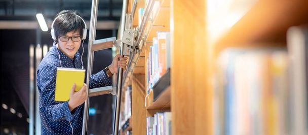 Azjata student uniwersytetu wybiera książkę z półki stojąc na drabinie w bibliotece do badań edukacyjnych. Zbiór bestsellerów w księgarni. Stypendium lub możliwości edukacyjne - Zdjęcie, obraz