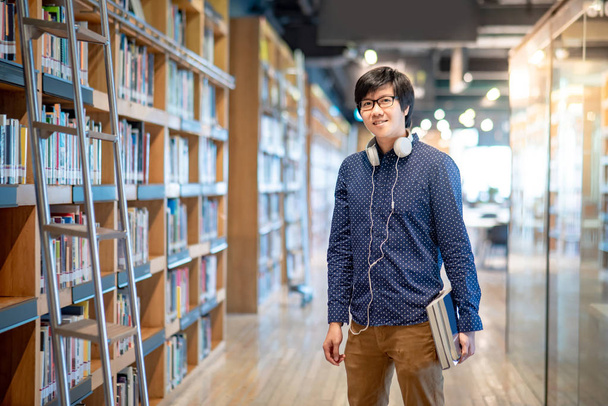 Ασιάτης φοιτητής που κρατάει βιβλίο κοντά σε ράφια βιβλιοθήκης σε βιβλιοθήκη κολλεγίου για εκπαιδευτική έρευνα. Συλλογή bestseller στο βιβλιοπωλείο. Υποτροφίες ή εκπαιδευτικές ευκαιρίες έννοιες - Φωτογραφία, εικόνα