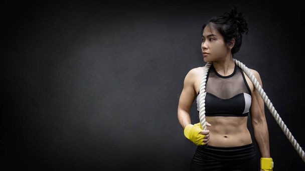 Asian girl fitness trainer posing with battle rope on shoulders on black background in studio. Physical exercise or workout in the gym. Muscular build or bodybuilding concepts - Photo, Image