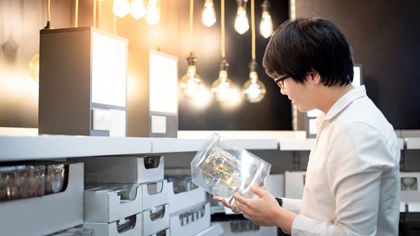 Азиатский парень покупатель выбирает пакет электрической лампочки с полки продукта на складе. Концепция покупки мебели для дома
 - Фото, изображение