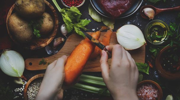 Womens hands are cleaning carrots. Cooking food background. Fresh organic vegetables, ingredients, spices and meat for soup on vintage kitchen table with rustic wooden cutting board. Top view. Healthy cooking and eating concept - 写真・画像