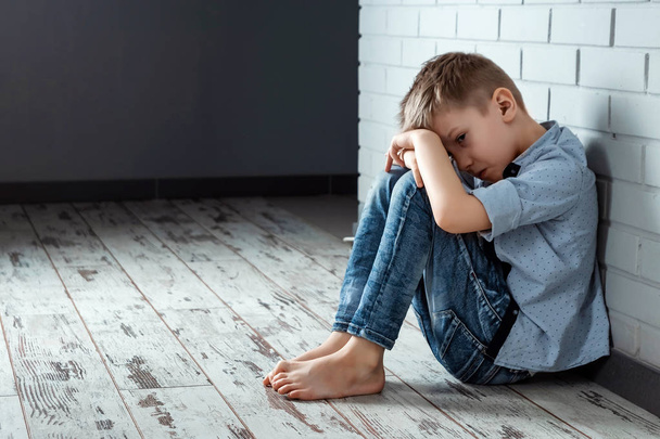 A young boy sits alone with a sad feeling at school near the wall. Offended child abandoned in the corridor and bent against a brick wall. Bullying, discrimination with copy space. - Фото, изображение