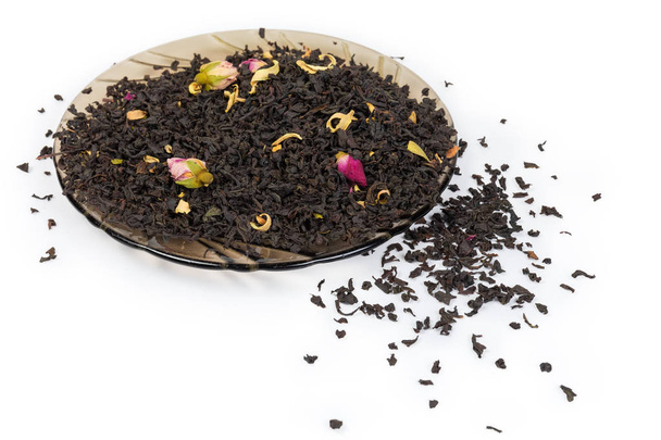 Folhas grandes secas de chá preto com aditação aromática de botões de rosa chineses e pétalas de flores de laranja no pires e espalhadas ao lado dele em um fundo branco
 - Foto, Imagem
