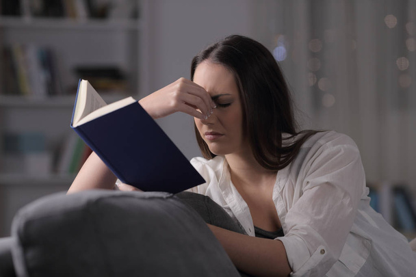 Dama que sufre fatiga visual leer libro sentado en un sofá en la noche en casa
 - Foto, imagen
