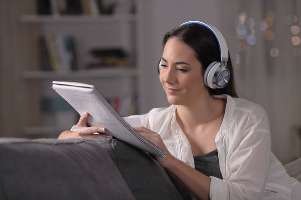 Serious student wearing headphones reading notes on a couch at home in the night - Фото, изображение