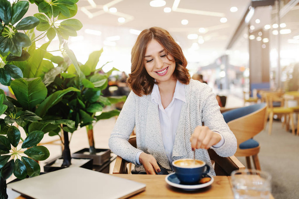 Charming brunette with toothy smile dressed casual stirring coffee while sitting in cafeteria. On desk glass of water and laptop. - Photo, Image