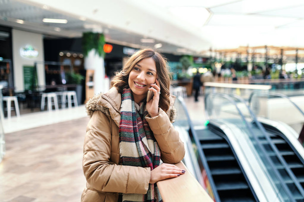 Uśmiechający się ładny brunetka w płaszczu i z szalikiem, opierając się na ogrodzenie i rozmawia inteligentny telefon stojąc w centrum handlowym. - Zdjęcie, obraz
