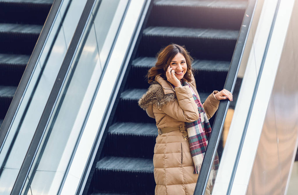 Smiling brunette in jacket and with scarf talking on the phone while riding escalator. Shopping mall interior. - Фото, изображение