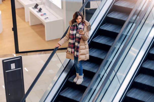 Vue du dessus de brune souriante en veste et avec écharpe parlant au téléphone tout en montant escalator. Intérieur du centre commercial
. - Photo, image