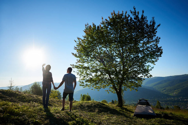 Back view of two happy young man and woman hikers resting near camping and big tree in mountains at sunny morning - Photo, Image