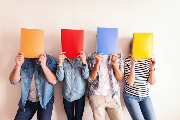 Four young business people standing against the white wall and holding folders in front of their faces. - Fotoğraf, Görsel