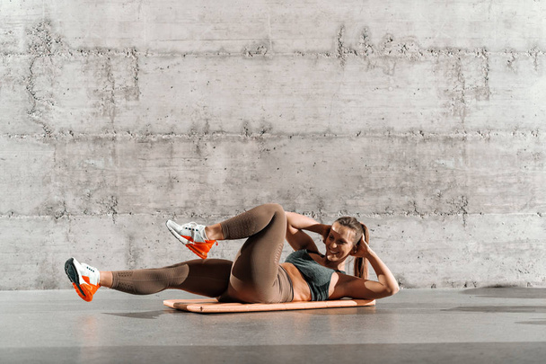 Lachende Kaukasische brunette in sport BH en legging doen abs liggend op de mat voor grijze muur in sportschool. - Foto, afbeelding