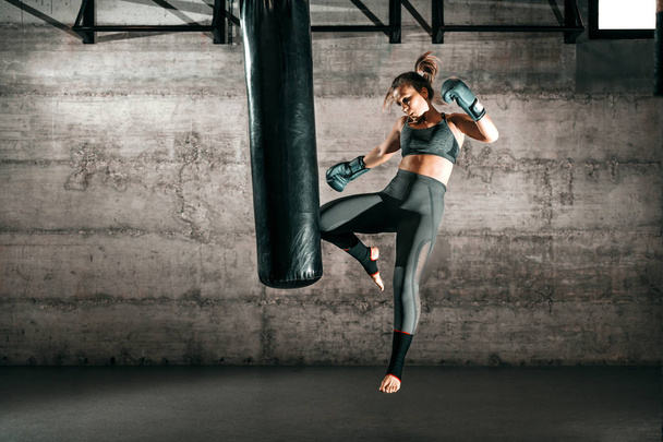 Dedicated strong brunette with ponytail, in sportswear, bare foot and with boxing gloves kicking sack in gym. - 写真・画像