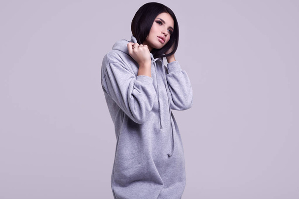 Portrait of gorgeous brunette woman in fashion blue hoodie posing on grey background in studio - Photo, image