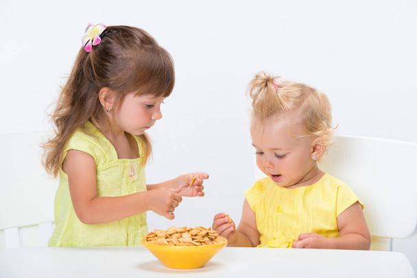 Two cute little girls sisters in yellow t-shirts eating cereal flakes at the table isolated on white background. - Photo, image