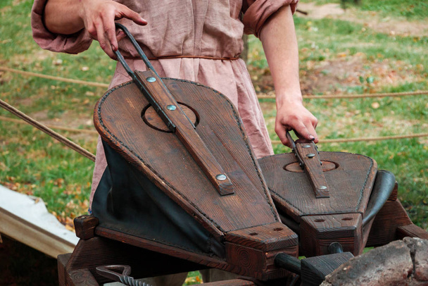 Blacksmith in ancient clothes squeezing bellows to kindle fire in furnace. Reconstruction - Foto, afbeelding