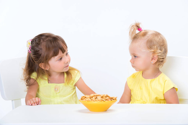Two cute little girls sisters in yellow t-shirts eating cereal flakes at the table isolated on white background. - 写真・画像