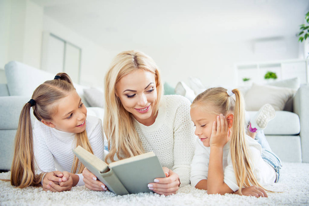 Portrait of three nice-looking cute pretty lovely sweet attractive charming cheerful people pre-teen girls mom mommy lying on carpet reading novel in light white interior room house indoors - Fotó, kép